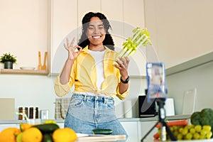 Food blogging. Happy black lady shooting on phone camera in kitchen, cooking and showing selera to smartphone on tripod