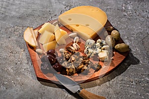 Food composition of cheese with dried olives, pieces of pear, pistachios and knife on cutting board on grey background.