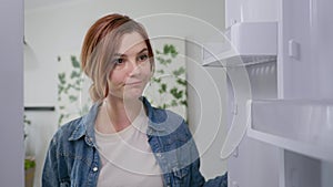 Food concept, young beautiful girl opens an empty refrigerator in search of meal looks sad because of lack of nutrition