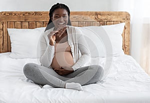 Food Cravings During Pregnancy. Cheerful Future Mom Eating Chocolate Bar In Bed