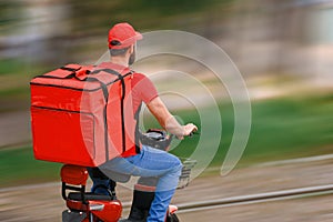 A food delivery man in a red uniform rides a moped with a food delivery bag.