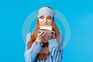 Food, eating and desserts concept. Cheerful pretty redhead woman in nightwear, sleep mask, close eyes smelling tasty