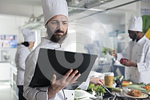 Food industry worker having laptop, following gourmet dish recipe on screen stirring ingredients in pan