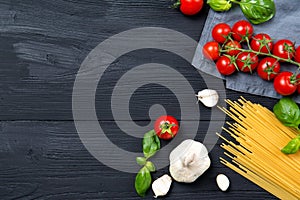Food ingredients for spaghetti pasta with cherry tomatoes, basil leaves and garlic, black wooden background, top view with copy