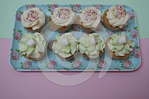 Food photography of a floral pattern tray filled with home made cupcakes with butter cream topping and a pink background