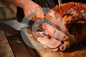 Food photography image of a cooked roast gammon or ham joint of meat with hand holding a carving knife and slicing the dinner
