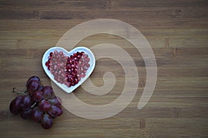 Food photography image of healthy red pomegranate seeds in a white love heart shape dish with purple grapes on wood background