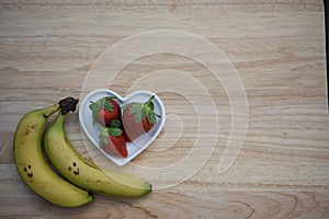 Food photography image of healthy red strawberries in a white love heart shape dish with bananas on wood background