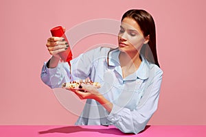 Food pop art photography. Young woman tasting hotdog with tomato ketchup isolated on pink background. Vintage, retro