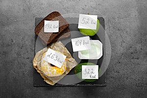 Food products with calorific value tags on dark grey table, flat lay. Weight loss concept