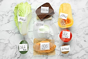 Food products with calorific value tags on white marble table, flat lay. Weight loss concept