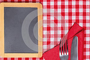 Food recipe template. Empty wooden black board and knife and fork on a red white checkered tablecloth. For your food and product