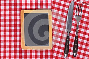 Food recipe template. Empty wooden black board and vintage knife and fork on a red white checkered tablecloth. For your food and