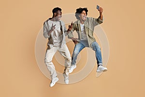 Fooling Together. Two Black Guys Taking Selfie While Jumping Over Beige Background photo