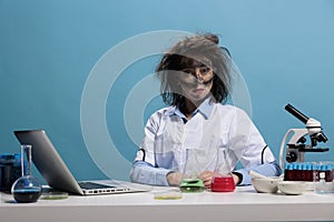 Foolish lab worker with wacky hair and dirty face sitting at desk on blue background while looking at camera.