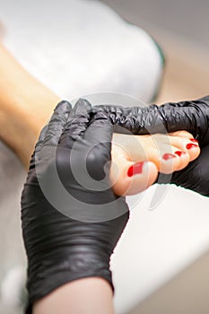 Foot massage with moisturizing and peeling cream by pedicurist hands wearing black gloves, close up.