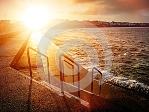 Foot path with metal barriers to the ocean and sunset sky over small town buildings. Kilkee city, county Clare, Ireland. Irish