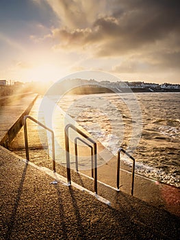 Foot path with metal barriers to the ocean and sunset sky over small town buildings. Kilkee city, county Clare, Ireland. Irish