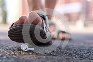 Foot in sandals steps on a lost wireless headphones, which lies on the asphalt sidewalk, outdoors, against a blurred