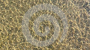 A footage taken from above the surface of a shallow, clear stream, revealing pebbles and sand with beautiful reflections.