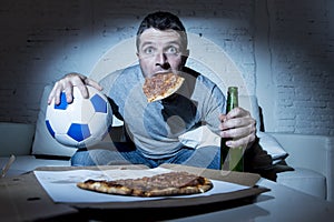 Football fan man watching soccer game on tv at home sofa couch with soccer ball and pizza in his mouth