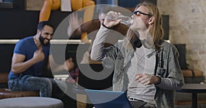 Football fans distracting concentrated young man using laptop and drinking water at front. Portrait of absorbed man