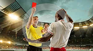 Football referee showing a player a red card for breaking rules at crowded stadium over night cloudy sky.