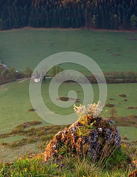 Podhorská krajina při východu slunce se skalami, loukami, stromy a starou architekturou v pozadí