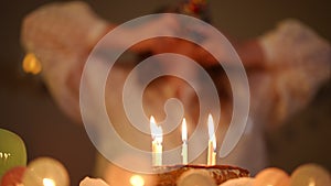 In the foreground is a birthday cake with burning candles. In the background, out of focus, a woman puts a birthday-cap