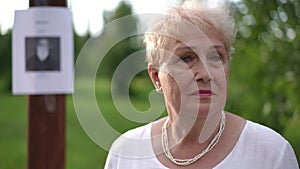 In the foreground, an elderly upset woman looks sadly at the camera while standing in a park outdoors on a summer day