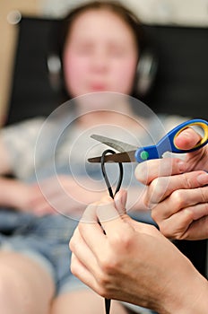 In the foreground, female hands are cutting the headphone cord with scissors, in the background is a girl in headphones