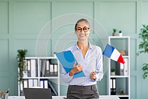 Foreign languages tutoring. Happy teacher holding flag of France and clipboard, standing near workdesk and smiling