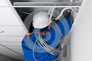 Foreman in blue uniform and white hard hat laying wiring for a Wi-Fi router in a hidden false ceiling system, close-up