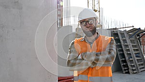 Foreman in safety helmet and vest stands at his workplace in a building under construction with his arms crossed in front of him