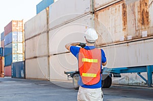 Foreman worker in hard hat and safety vest talks on two-way radio control loading containers box from cargo