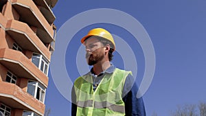 Foreman in a yellow helmet with a beard and mustache goes near the house under construction and tells about the construction proce