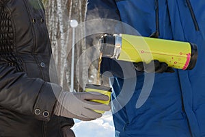 In the forest, in the cold, a man pours a woman hot tea from a thermos