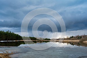 Forest lake with ice remnants, trees on the shore and dark rainy clouds in the sky. Wildlife spring landscape on cloudy evening