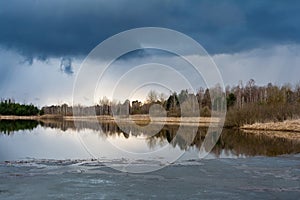 Forest lake with ice remnants, trees on the shore and dark rainy clouds in the sky. Wildlife spring landscape on cloudy evening