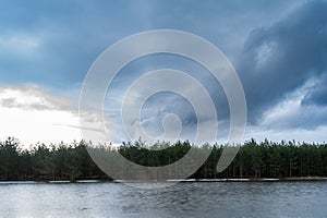 Forest lake with ice remnants, trees on the shore and dark rainy clouds in the sky. Wildlife spring landscape on cloudy evening