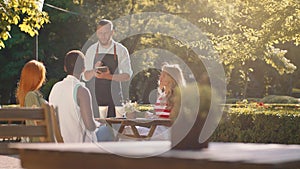 In a forest scenery, a very attractive waiter with a beard and hair tied back is taking orders from a table with three