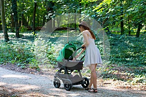 Forest walk with stroller, side view of young mom in beautiful dress walking on the pathway with her infant in the pram