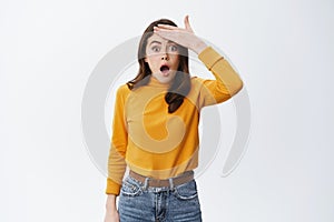 Forget remember. Shocked woman slap forehead and gasping startled at camera, standing in casual yellow sweater and jeans