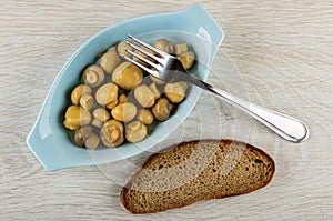 Fork on blue plate with marinated champignons, slice of bread on wooden table. Top view