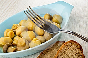 Fork on blue plate with marinated champignons, slices of bread on wooden table