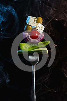 Fork with ingredients for traditional greek salad with fresh vegetables, feta cheese and olives on black background