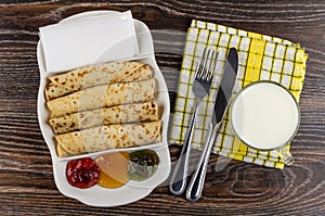 Fork and knife, cup of milk on napkin, dish with jams, pancake rolls, tissue on table. Top view