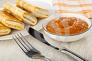 Fork and knife, plate with fried pancake rolls, napkin, bowl with squash caviar on table