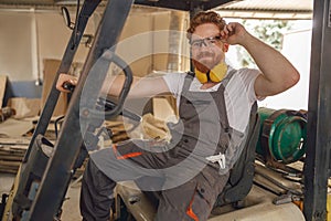 Forklift driver in protective eyeglasses sitting in vehicle in wood warehouse and looks camera