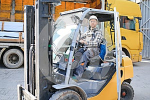 forklift driver in protective vest and forklift standing at warehouse of freight forwarding company, smiling